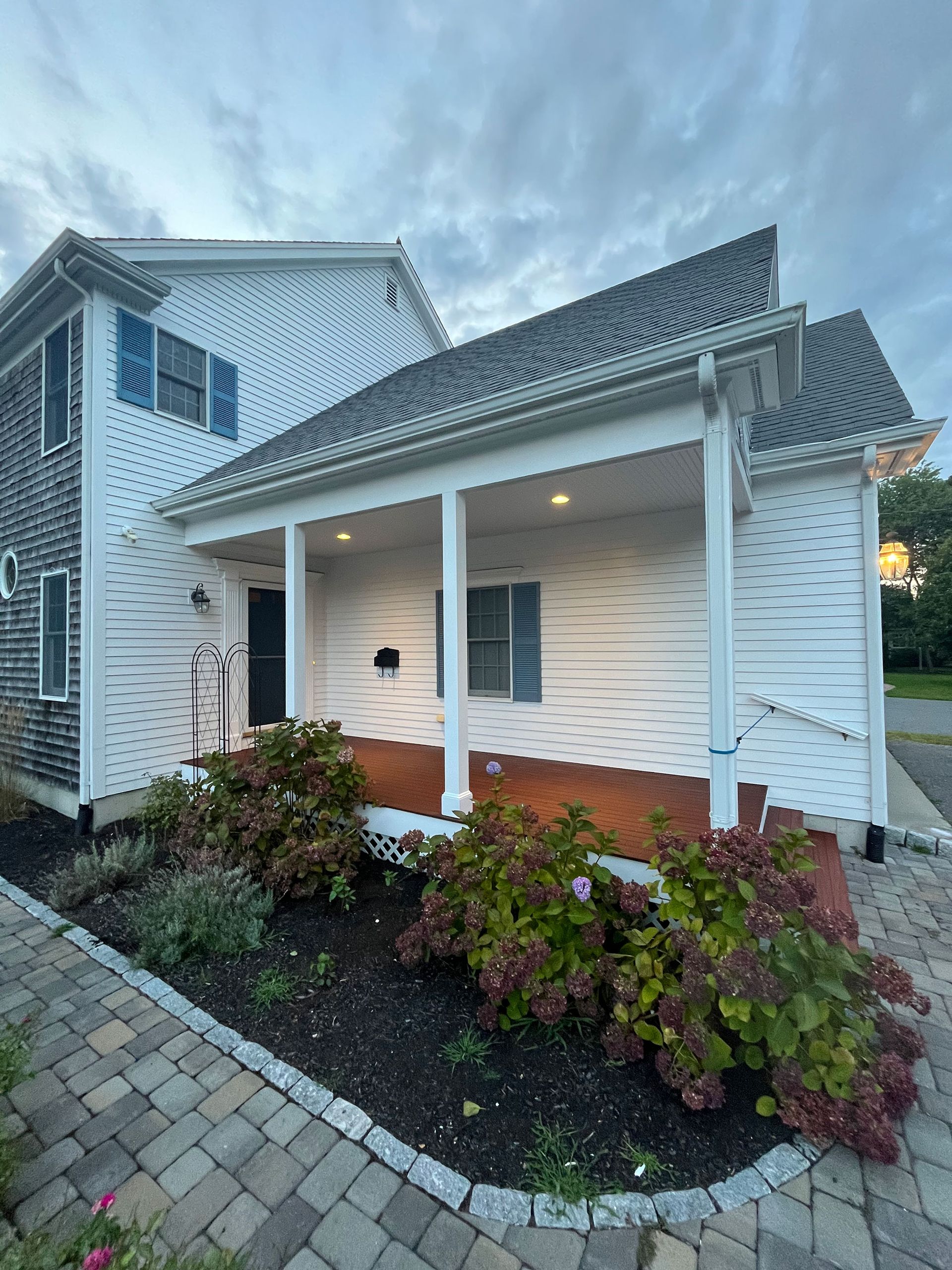 Two-story house with white siding and a covered porch, plants, and a brick walkway.