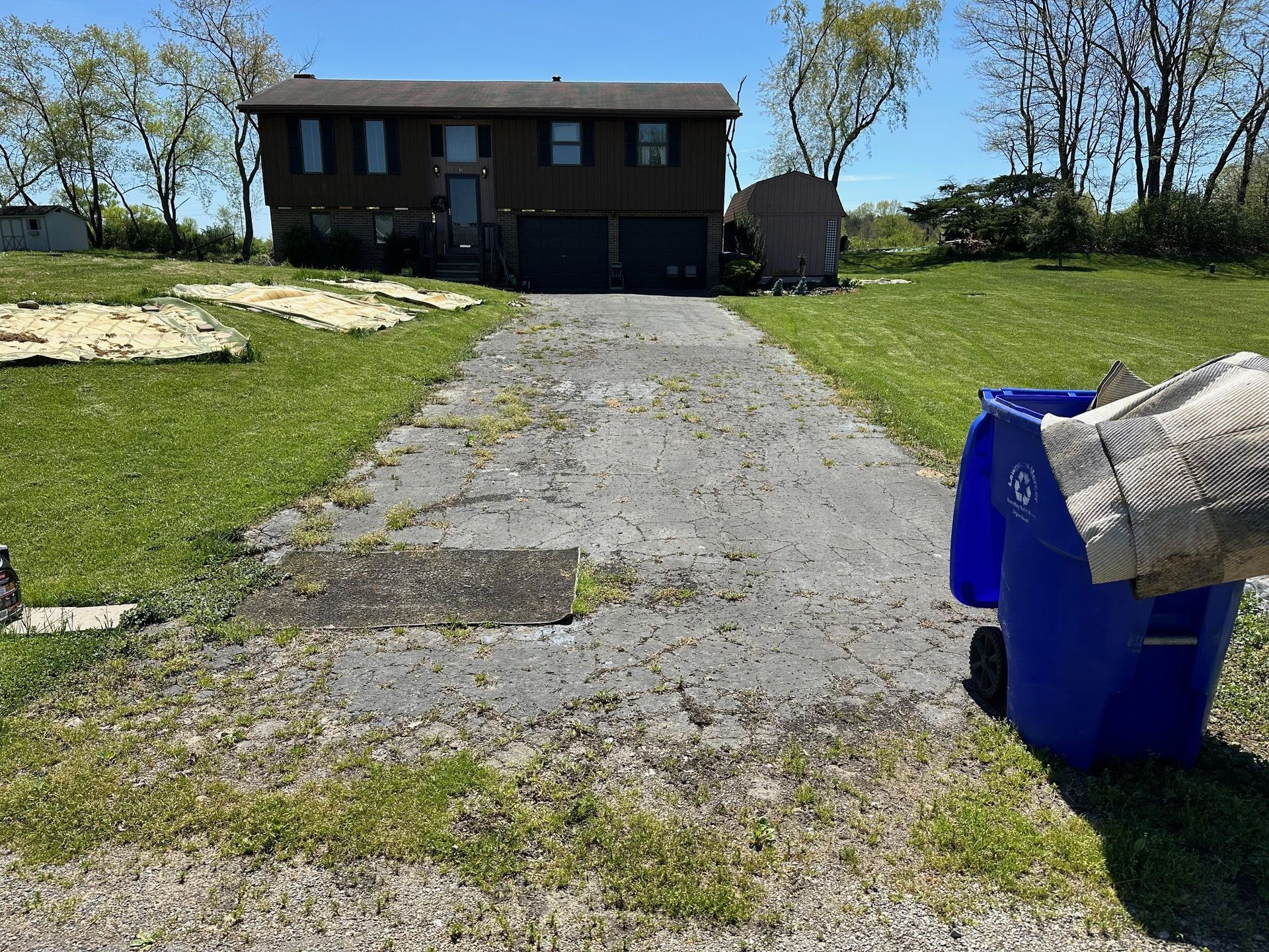 A blue trash can is sitting on the side of a dirt road in front of a house.