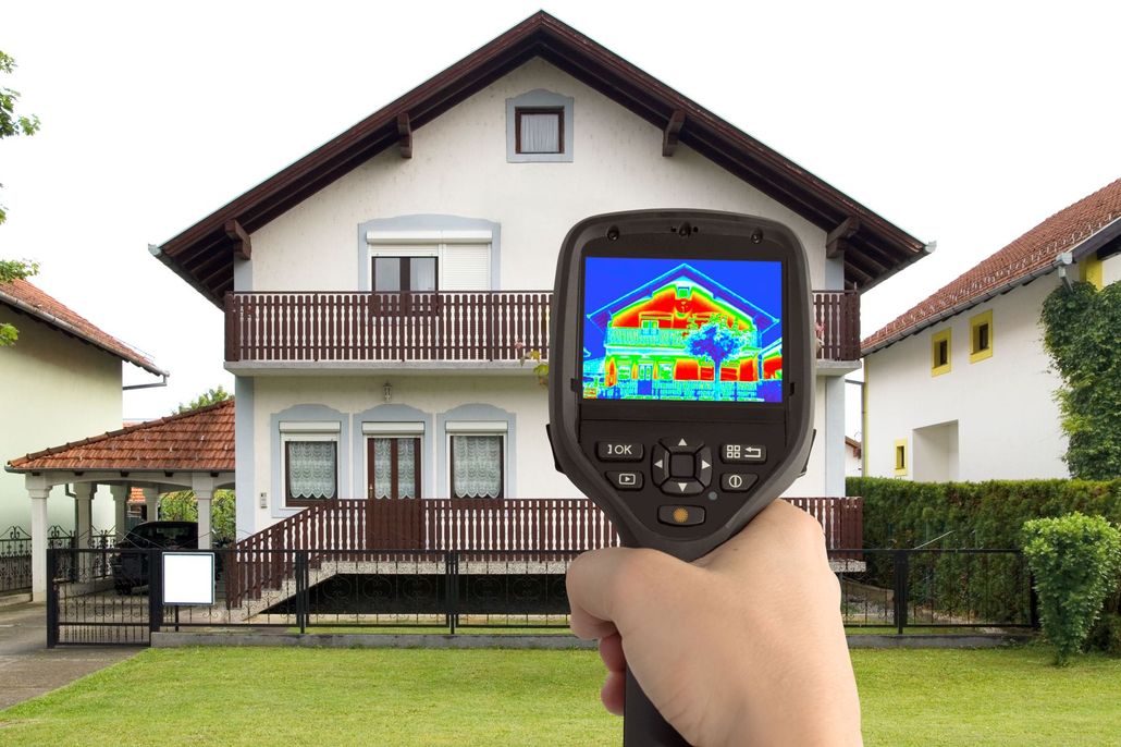 Hand holding thermal camera pointed at house, showing heat loss in red/yellow colors.