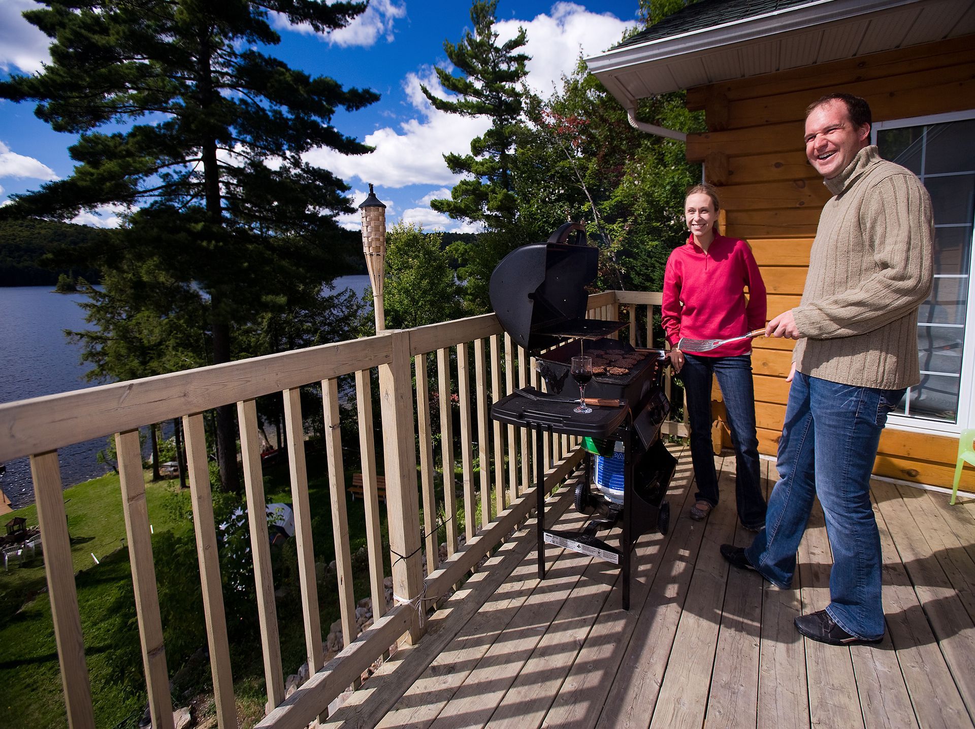 A man and a woman are grilling on a deck overlooking a lake.