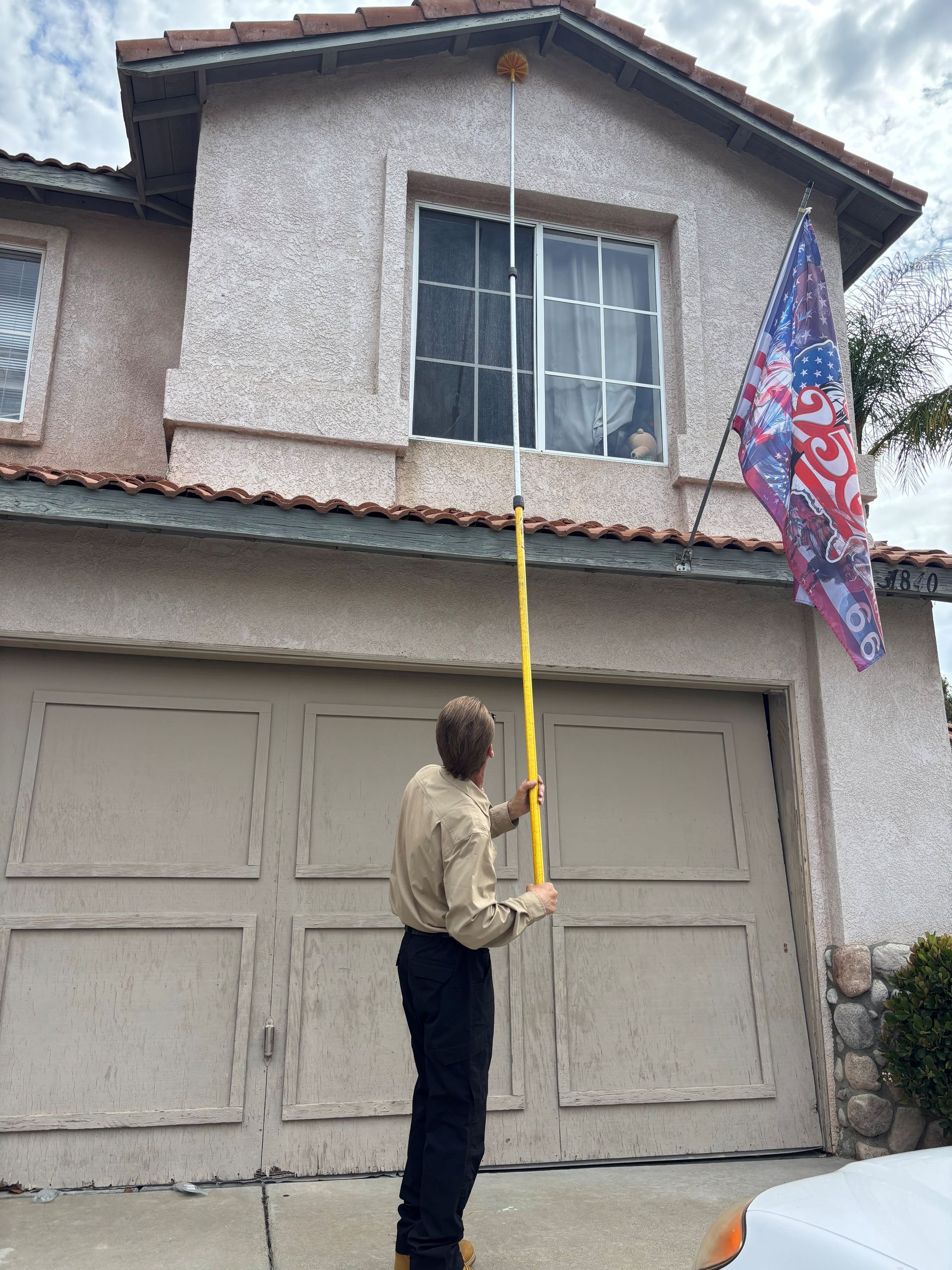Person on a driveway using a long yellow pole to kill spiders and remove webs and spider eggs on a two-story house