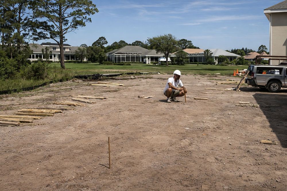 Construction worker on dirt lot, inspecting with level; houses in background.