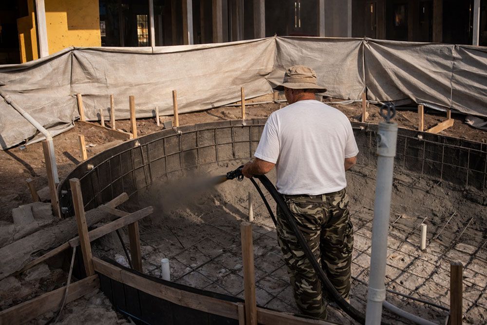 Man spraying concrete into a pool's framework during construction. Outdoors, wearing a hat, white shirt, and camo pants.