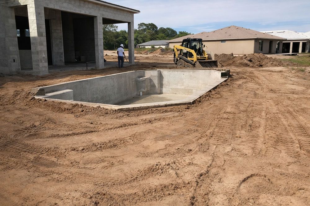 Construction site with a concrete pool, a small excavator, and a person near a partially built house.