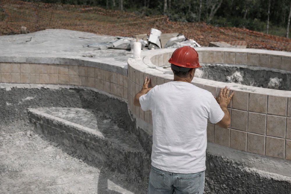 Construction worker in red hard hat installing tiles on curved pool structure.