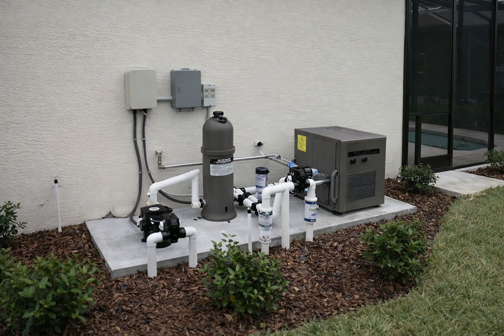 Pool equipment set against a beige wall, including a filter, pump, heater, and control boxes on a concrete pad with landscaping.