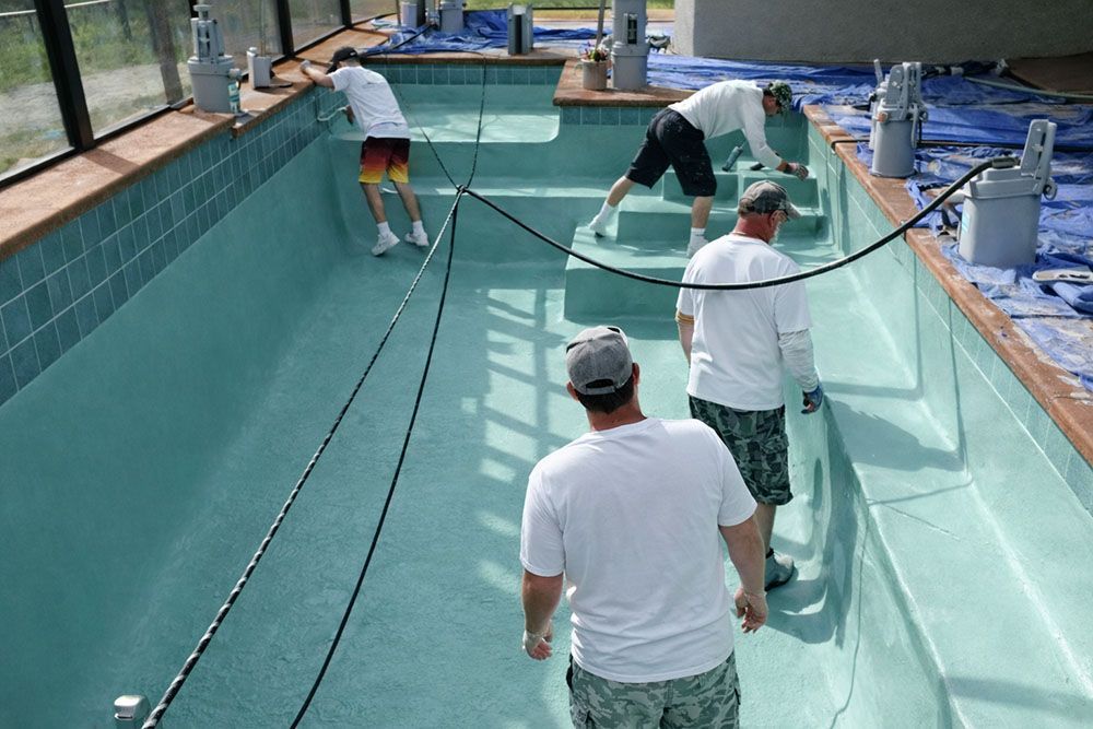 Four people working inside an empty, turquoise-tiled swimming pool. They wear light-colored shirts, shorts, and hats.