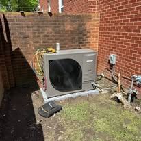 An air conditioning unit sits against a brick wall. The unit is gray, on a concrete base.