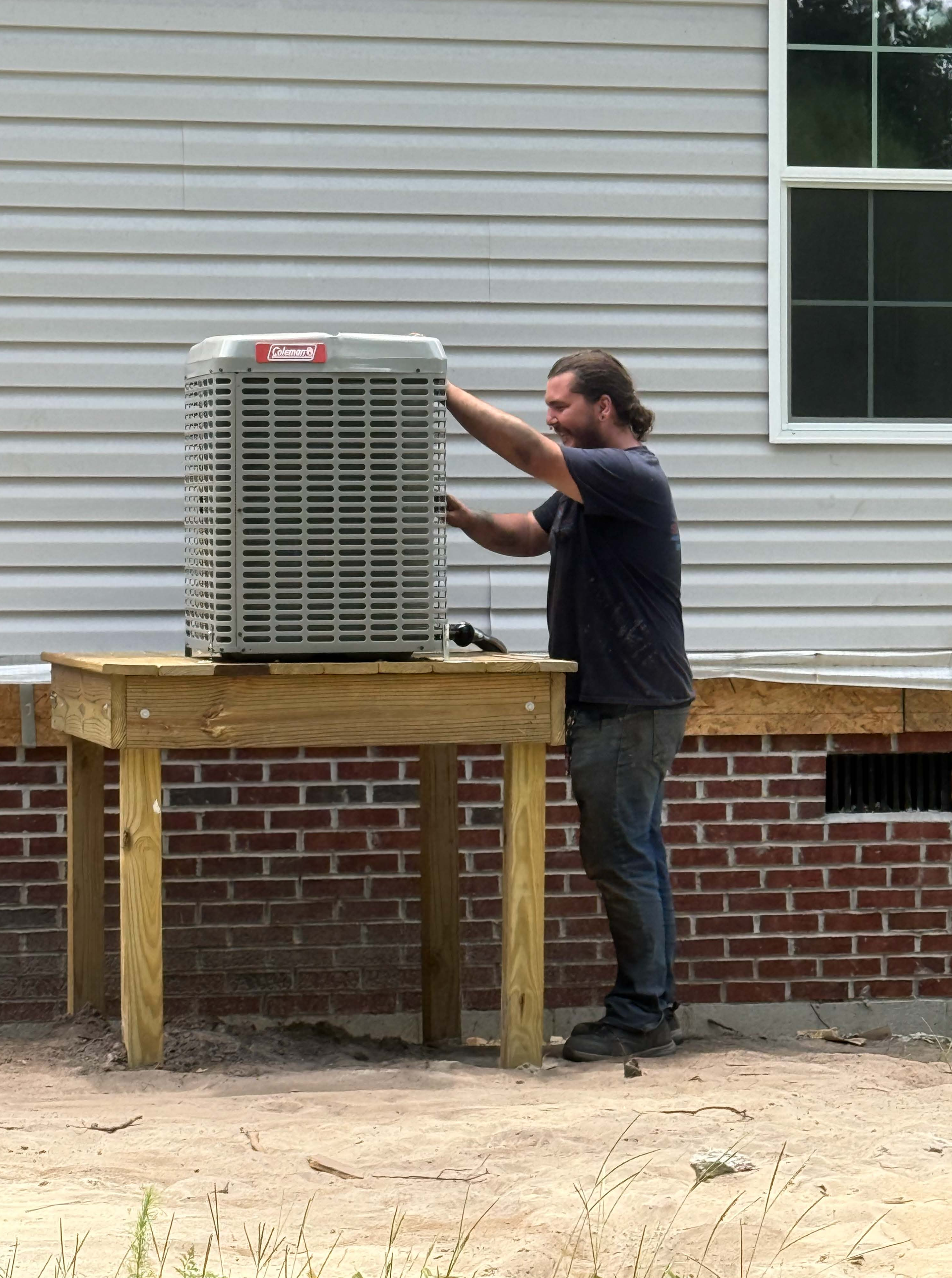 Man installing a new air conditioner on a wooden platform next to a house with brick and siding.