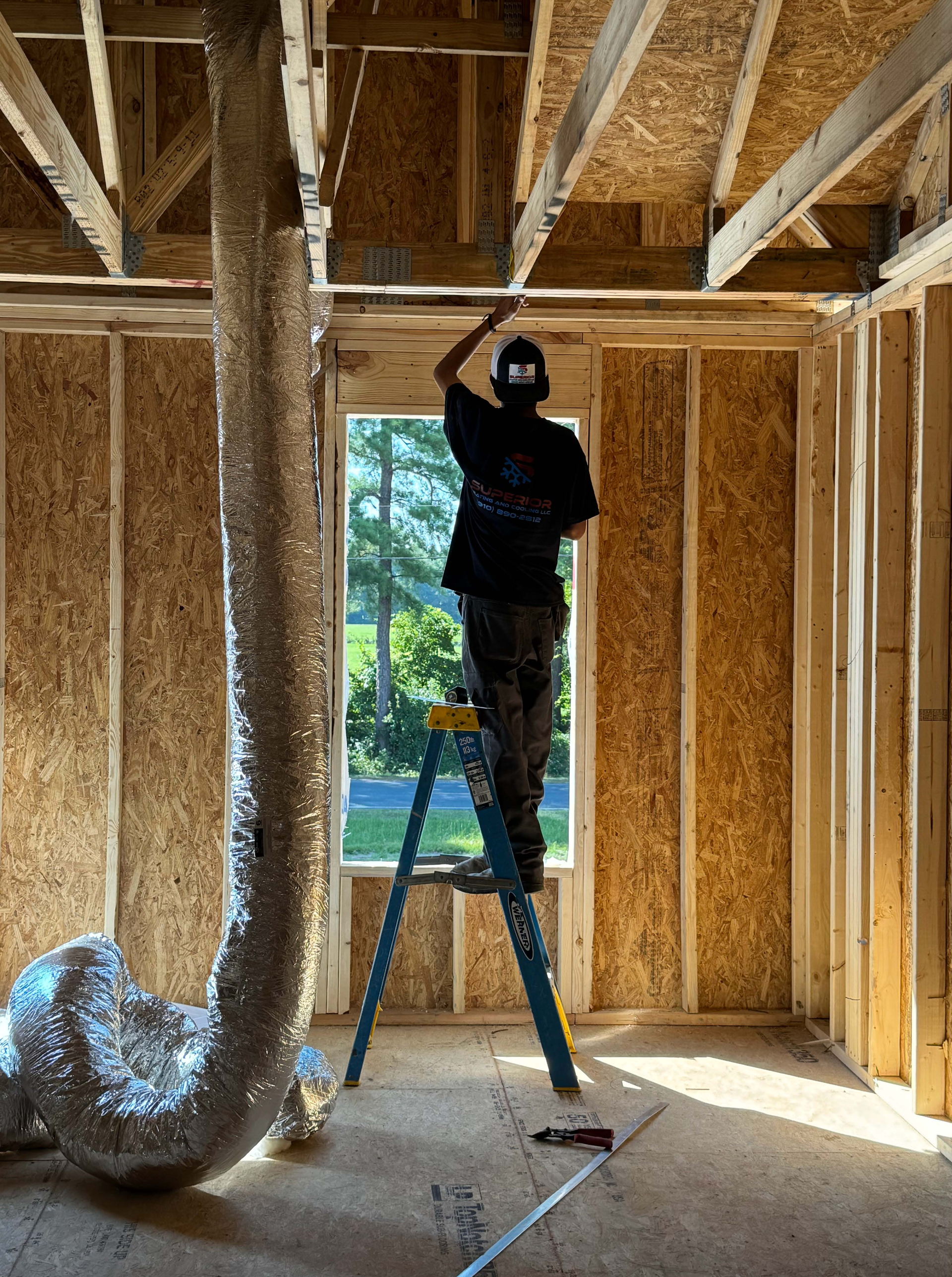 A person on a ladder installing ductwork in a house under construction.