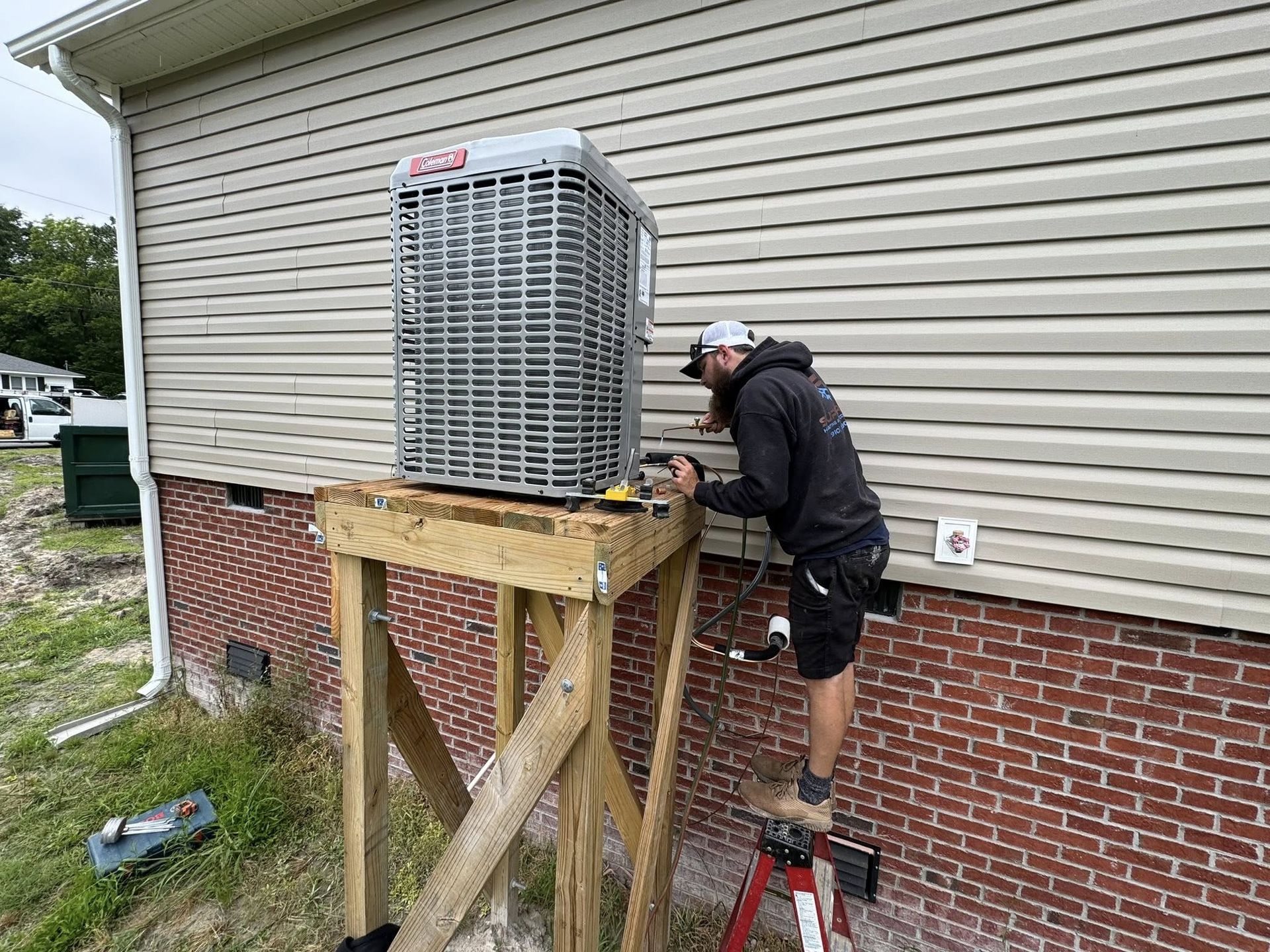 Man installs an AC unit on a wooden stand near a brick foundation of a house.
