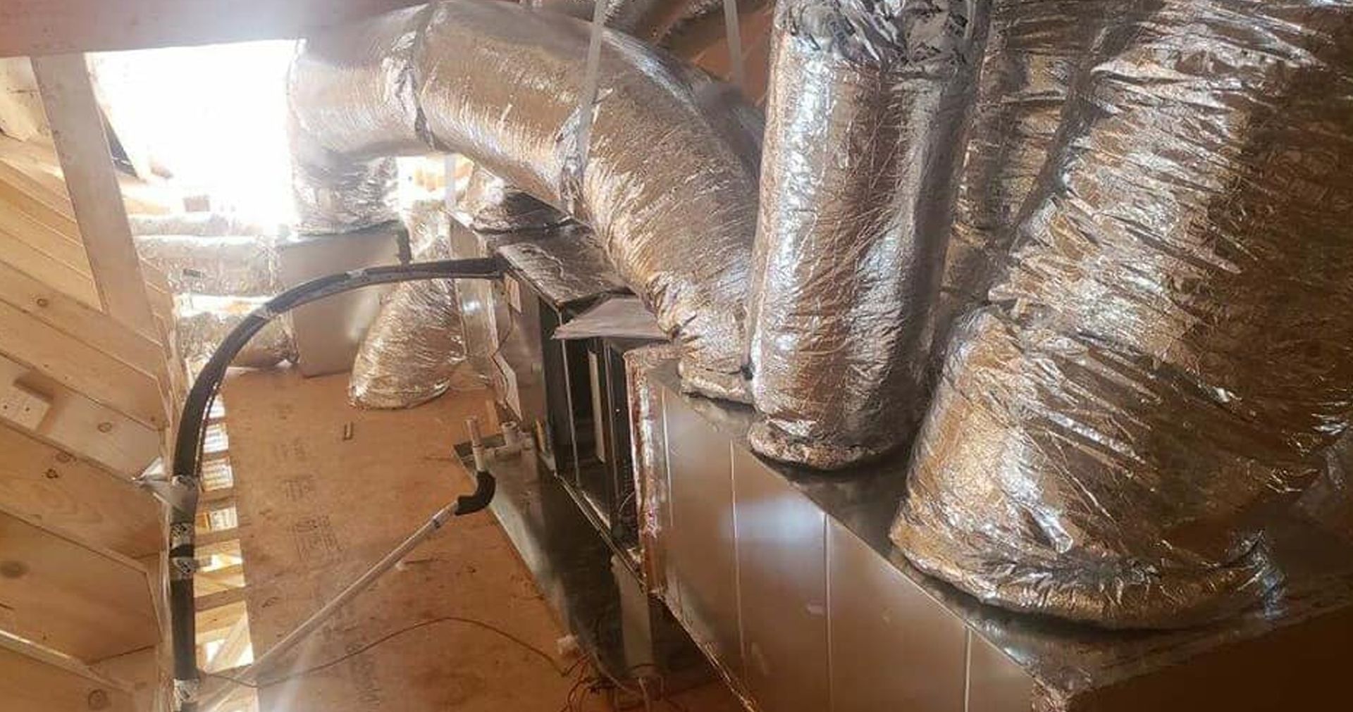 Man installing flexible silver ductwork in a ceiling cavity. He is wearing a white shirt. The setting is an unfinished interior space.