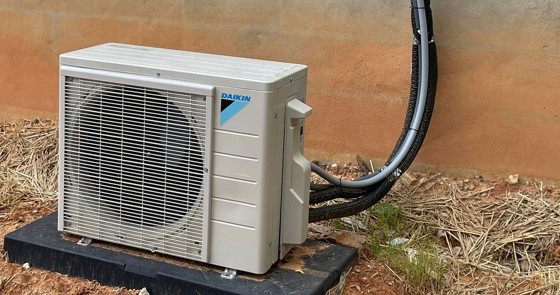 Exterior view of a light grey mini-split air conditioning unit mounted on a textured brown wall, with a power cord and electrical box.