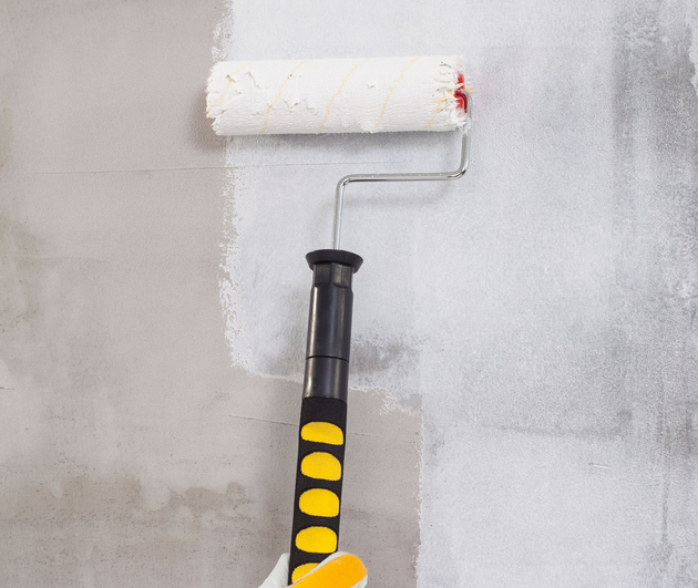 Gloved hand painting a wall with a roller, covering a gray surface with white paint