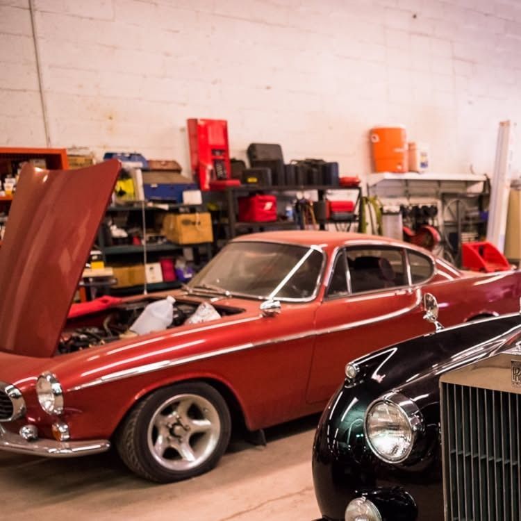 Red classic car in a garage with shelves of tools and parts, next to a black vintage car.
