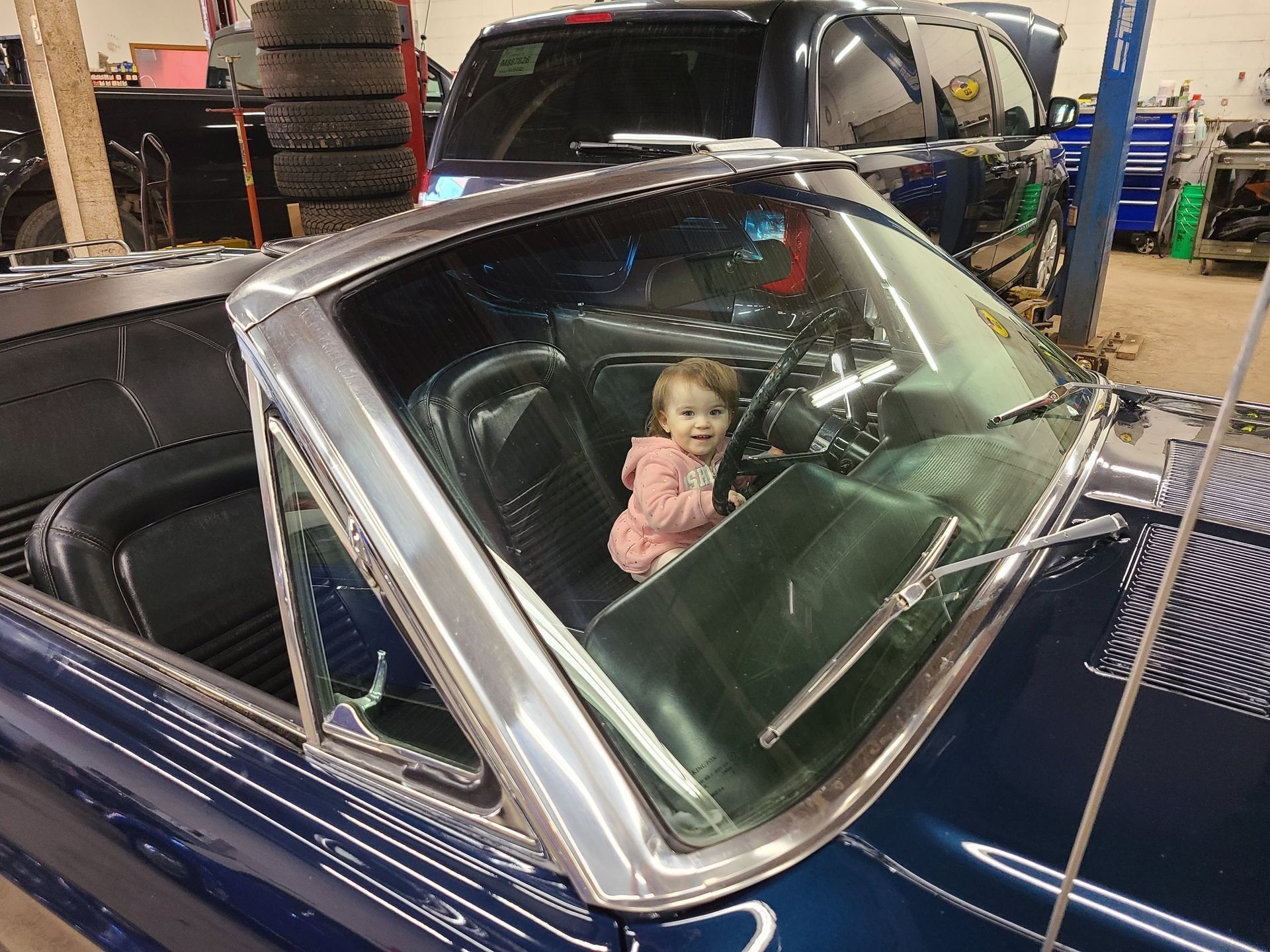 Toddler sitting in the front seat of a blue convertible in a garage, smiling through the windshield