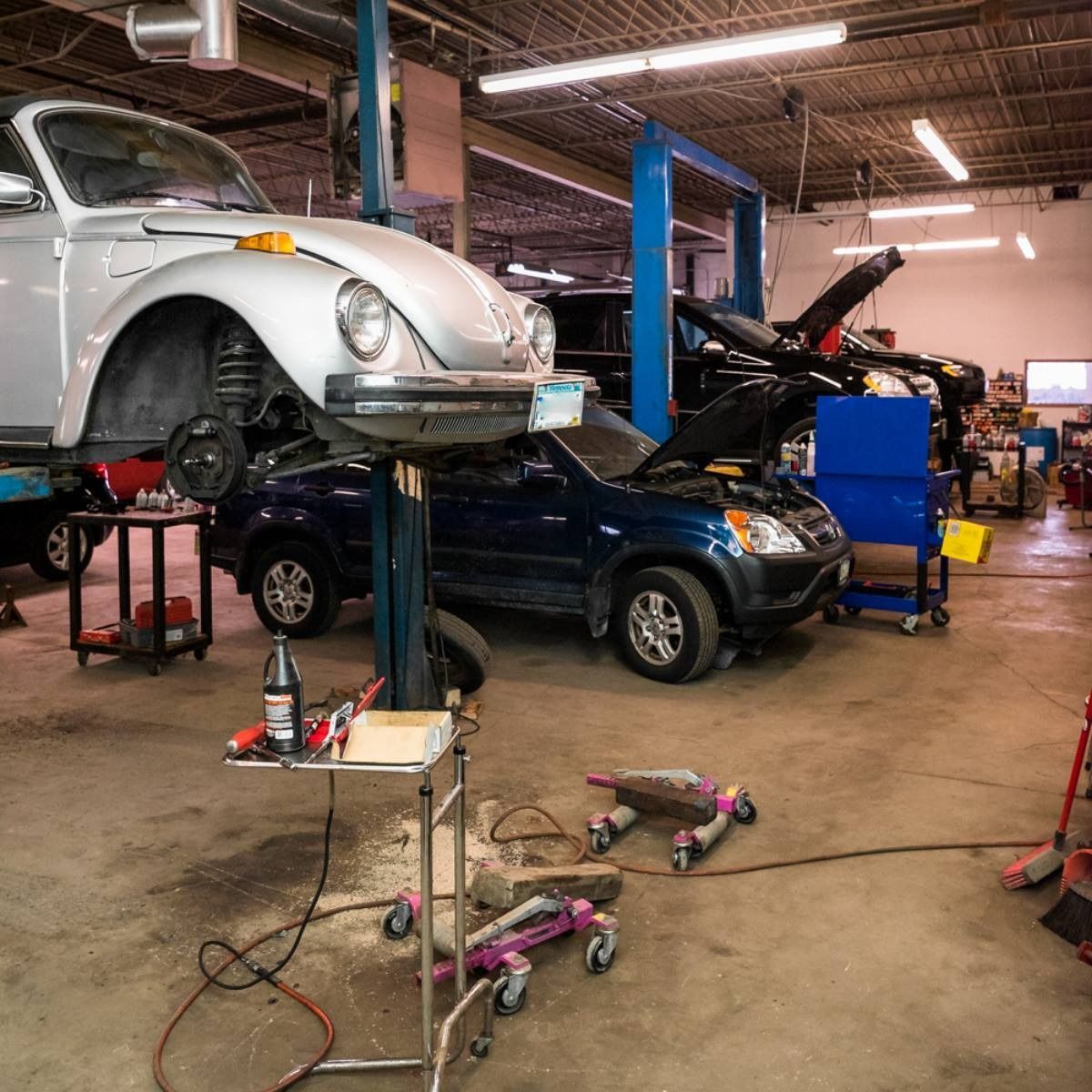 Auto repair shop with a white VW Beetle on a lift over a dark blue car, tools scattered on the floor.