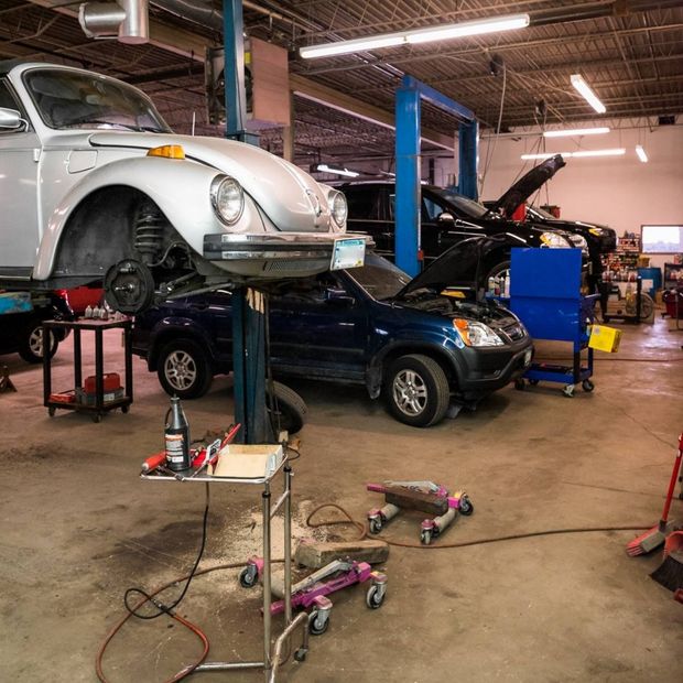 Auto repair shop with a white VW Beetle on a lift over a dark blue car, tools scattered on the floor.
