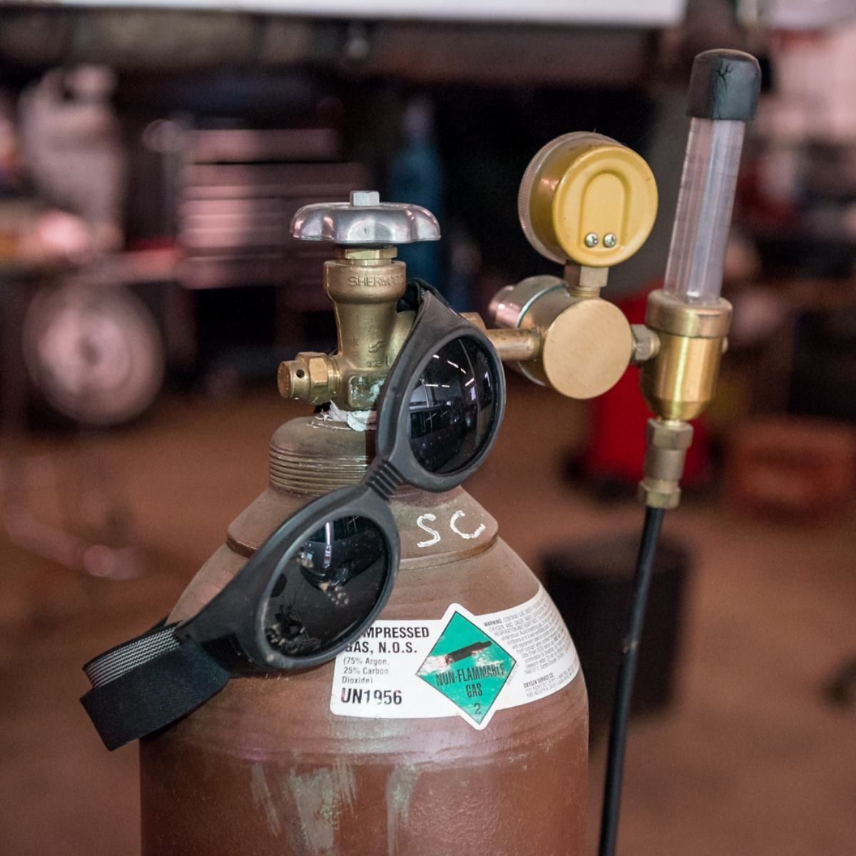 Brown gas cylinder with attached regulator, gauges, and black safety glasses in a workshop setting