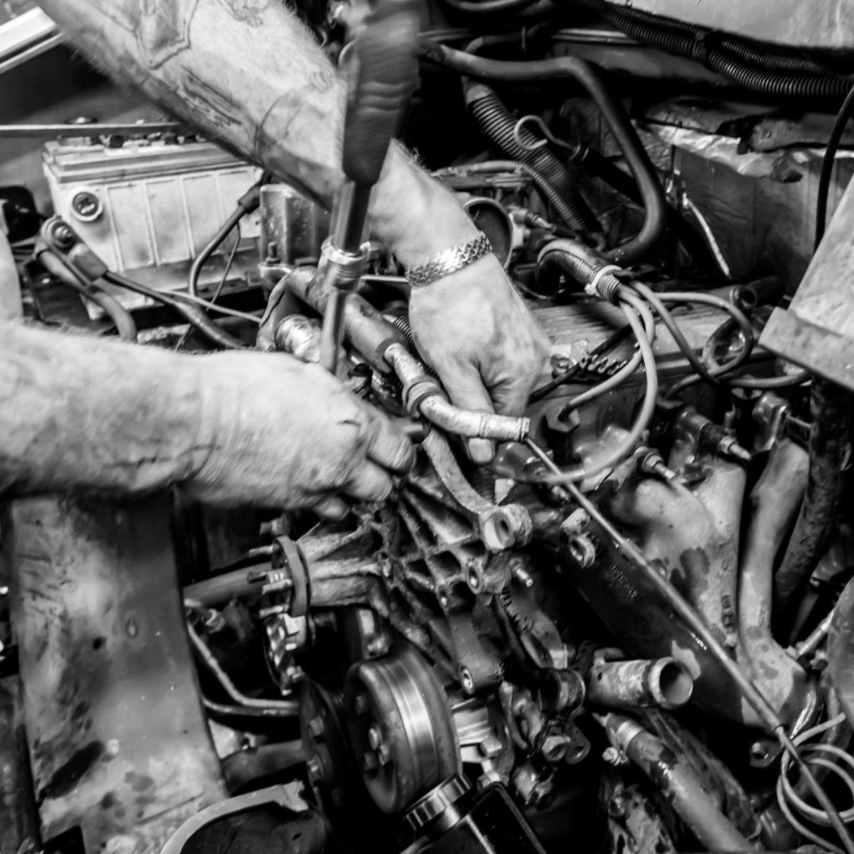 Mechanics repairing a car engine, with hands working amid exposed hoses and parts.