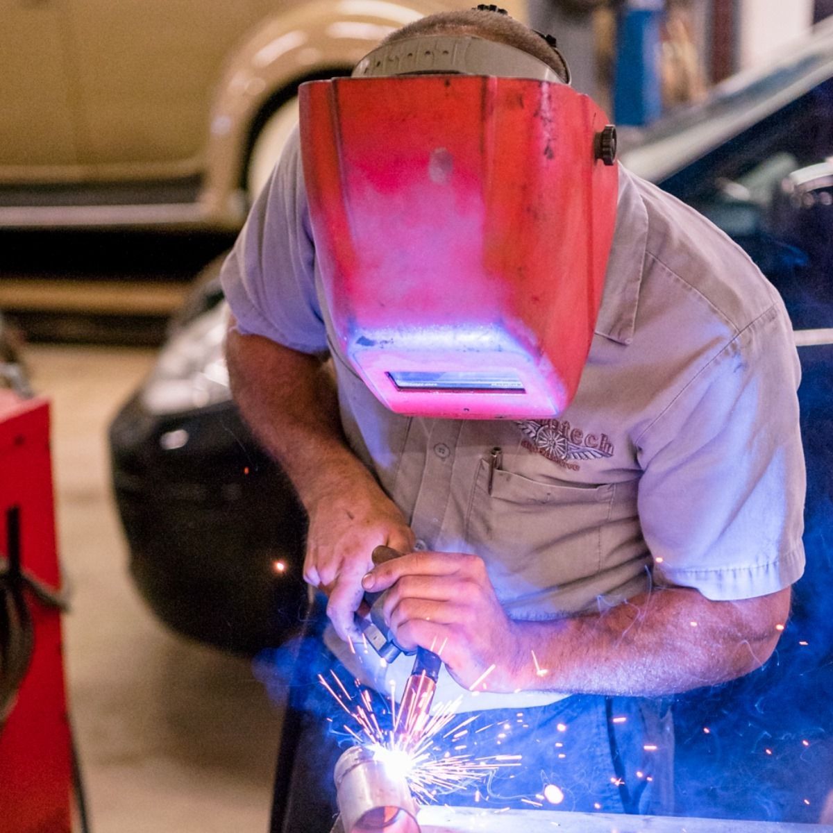 Person welding metal indoors, wearing a red helmet and white shirt, with bright sparks flying.