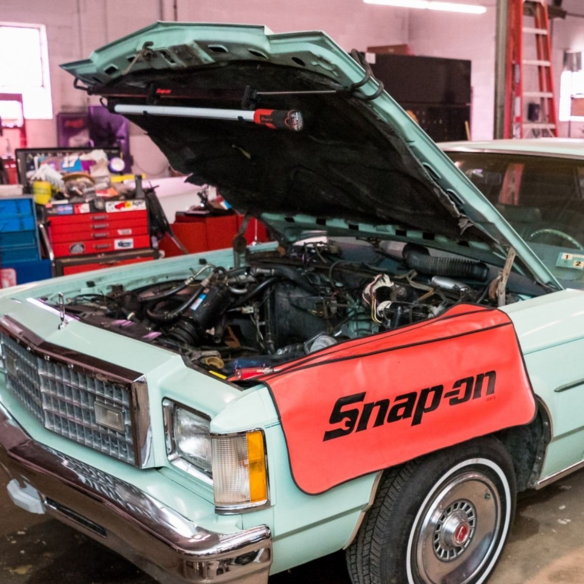 Mint-green vintage car with hood open in a mechanic’s shop, featuring a red Snap-on fender cover