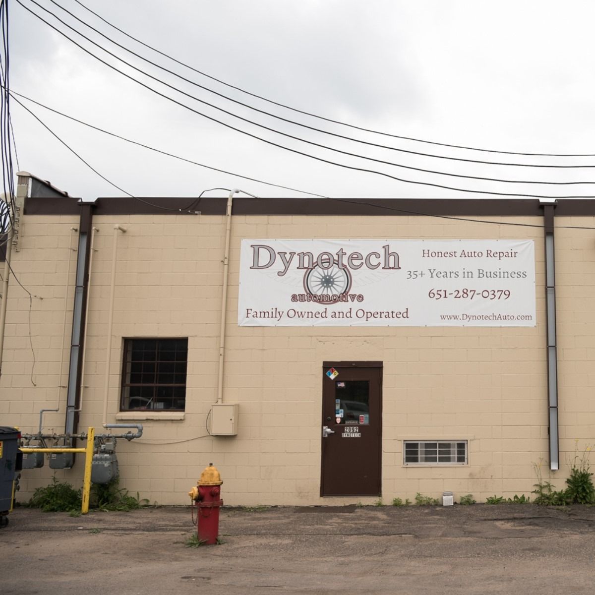 Beige Dynatech building with sign, door, windows, fire hydrant, and overhead power lines.