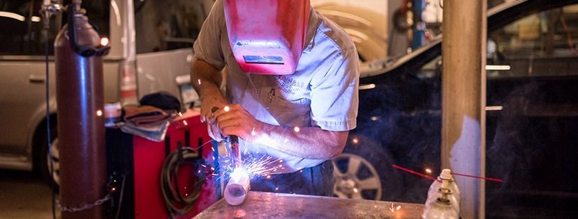 Welder in a mask sparks metal in a workshop with welding equipment and tools around.