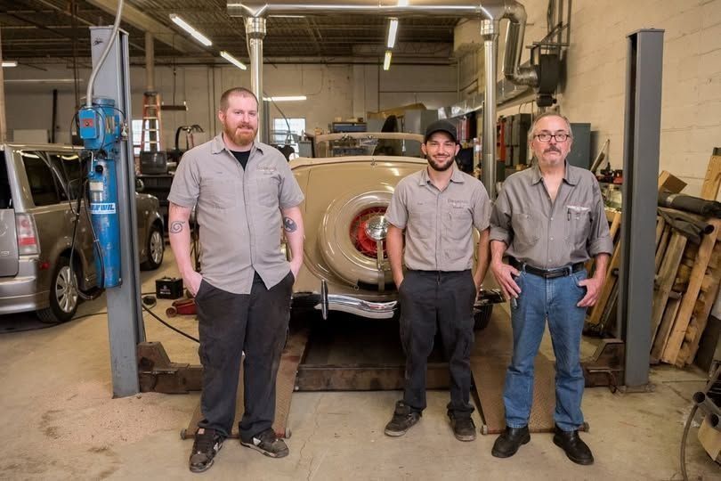 Three mechanics stand in a garage with a car on a lift behind them.