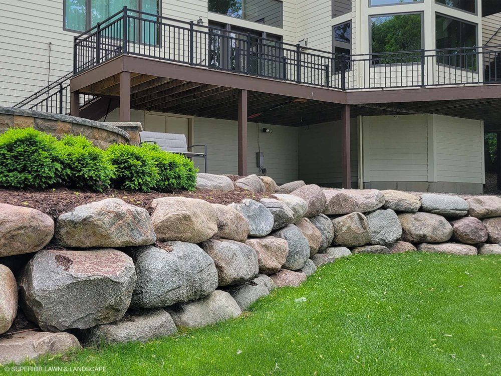 A large rock wall is in front of a house with a deck.