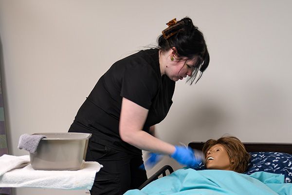 Person in blue gloves washes the face of a mannequin lying in a bed. A basin of water and washcloth is nearby.