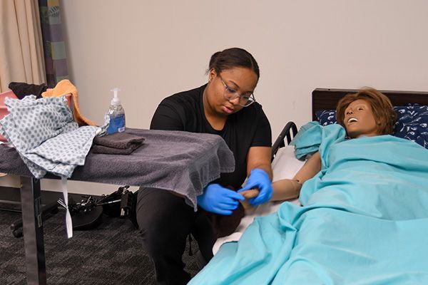 Person in scrubs tending to a mannequin patient in a simulated hospital room.