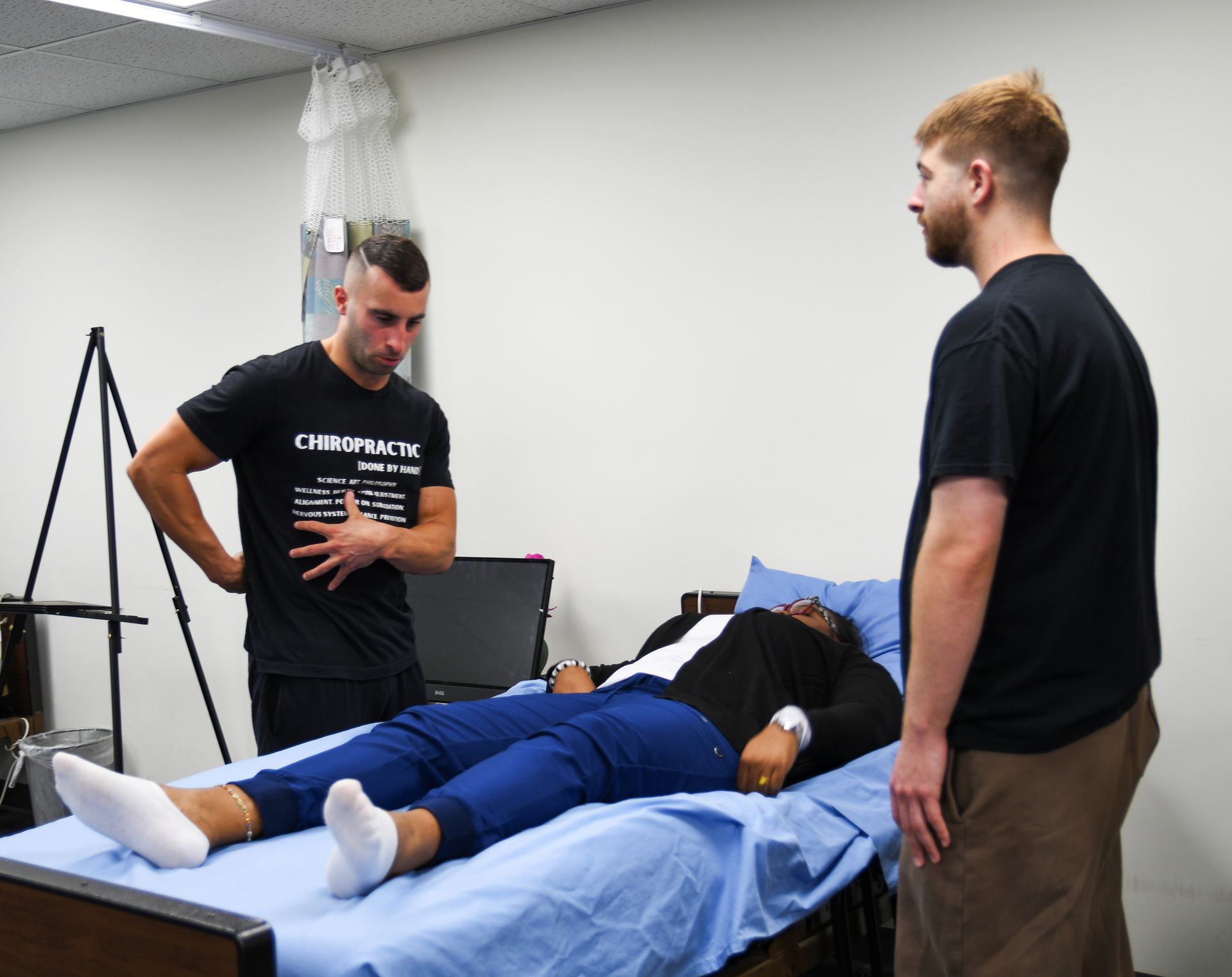 Three people in a medical setting. One lies on a bed, while two others stand near, examining and talking.