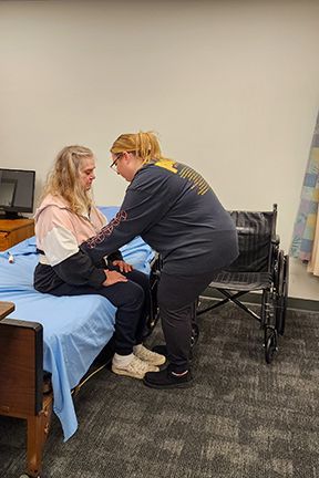 A person in casual clothes assists another person from a bed into a wheelchair.