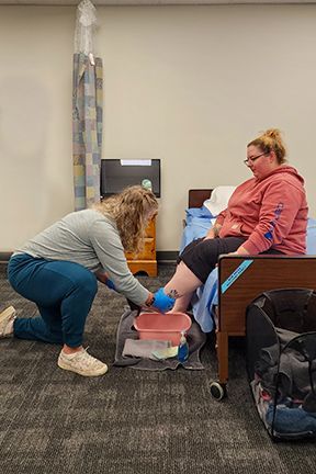 Person kneels, washing another's foot in a basin. Both are indoors, near a bed and equipment.