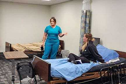 Nurse in teal scrubs talks to a woman sitting on a hospital bed; clinical setting.