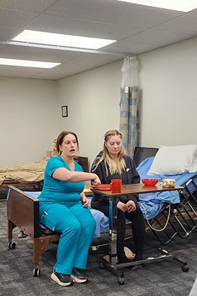 Woman in teal scrubs points at object; woman sits, a bed in background.