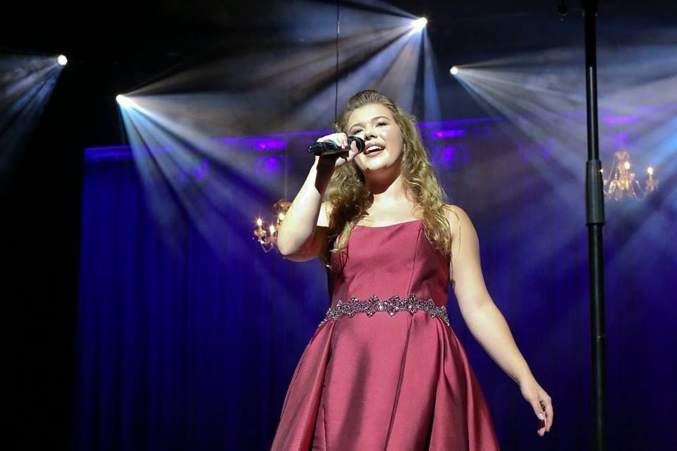 Woman in burgundy dress singing on stage, lit by spotlights.