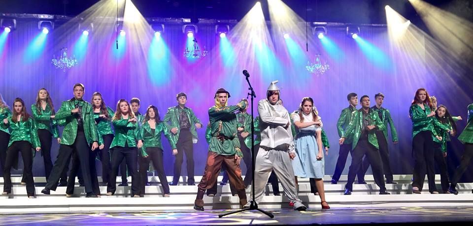 Musical theater performance on stage with ensemble in green jackets, and featured characters in costume.