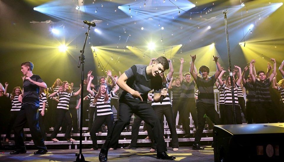 A choir performs on stage with a spotlight. One singer dances as the others sing with arms raised.