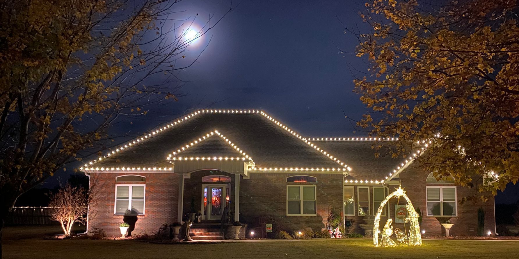 A house at night, decorated with white Christmas lights on the roof and tree. A bright full moon in the dark sky.