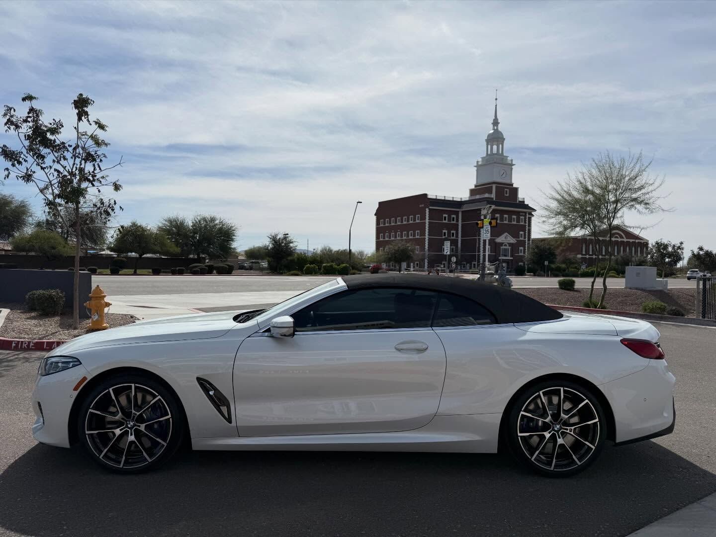 White sports coupe parked outdoors in front of a brick building with a tall steeple under a cloudy sky