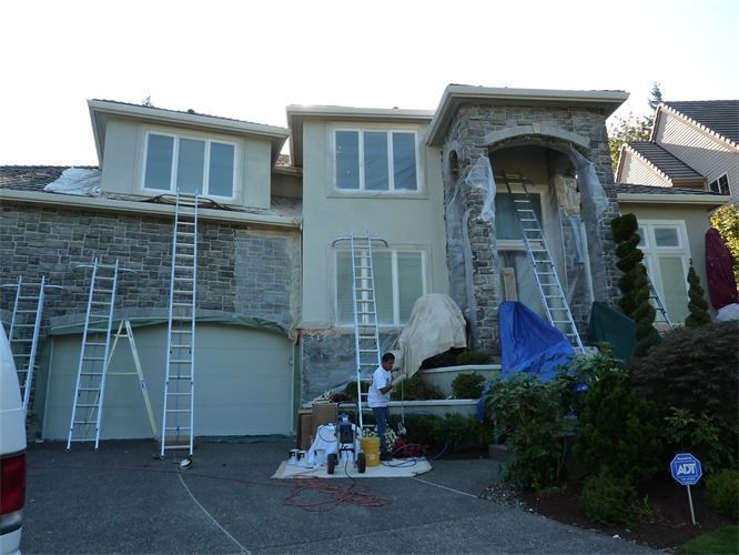 Exterior house being painted; workers on ladders. Beige stucco, stone facade, ladders, blue tarp, paint supplies.