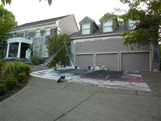House exterior being painted, with ladders, tarp, and paint cans. Driveway in foreground.