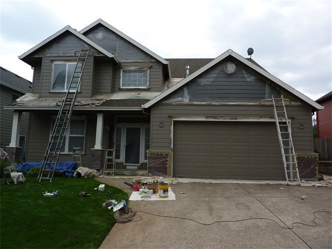 House exterior under renovation: Ladders, siding partially removed, brown paint, tools on ground, gray sky.