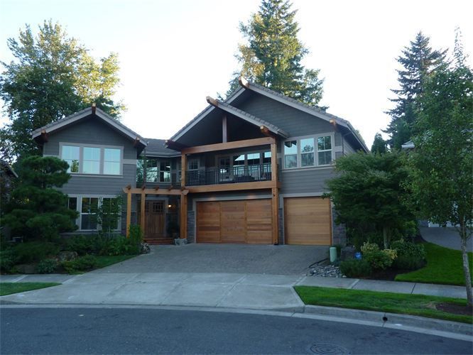 Two-story gray house with wooden accents, two-car garage, balcony, and driveway. Green grass and trees surround it.