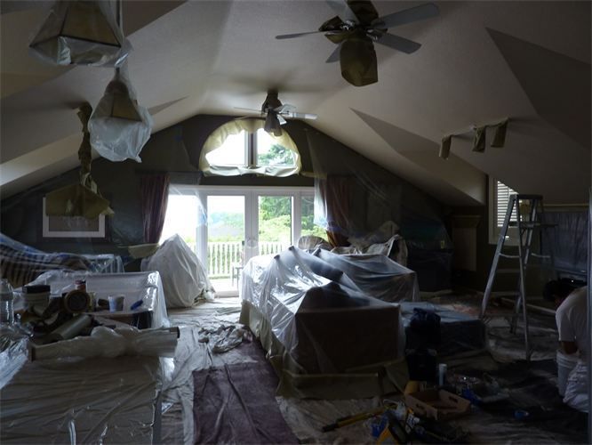 Room under renovation, covered furniture, painter, protective plastic, vaulted ceiling, windows, natural light.