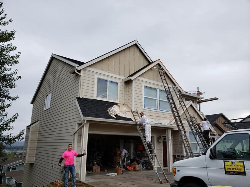House exterior being painted; men on ladders, beige and brown siding, gray sky.