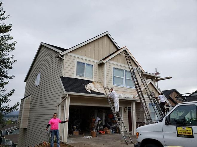 House exterior being painted; men on ladders, beige and brown siding, gray sky.