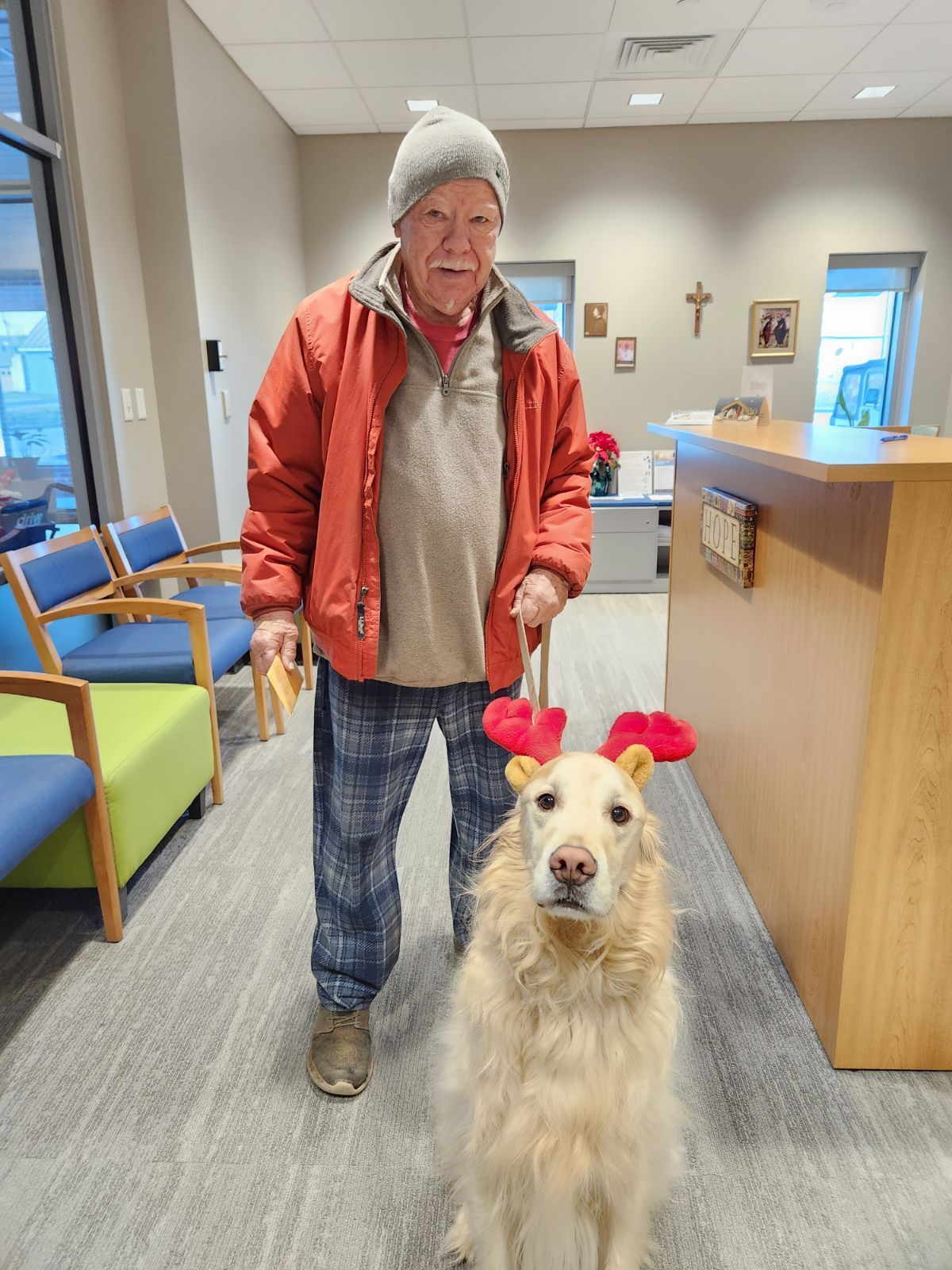 Older man in winter coat standing with light brown long-hair golden retriever dog wearing reindeer antlers.